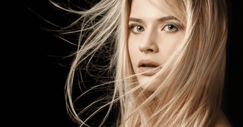 Retrato de mulher com cabelos longos e loiros em diferentes nuances claras, com fios em movimento sobre o rosto e fundo preto.