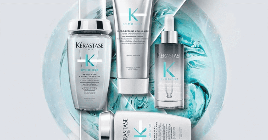 Produtos da linha Kérastase Symbiose dispostos sobre fundo azul translúcido, incluindo shampoo, micropeeling, sérum e tratamento para couro cabeludo e cabelos.