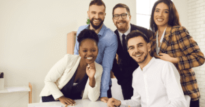 Grupo de cinco profissionais sorrindo em ambiente de escritório, vestindo roupas de trabalho, posados ao redor de uma mesa
